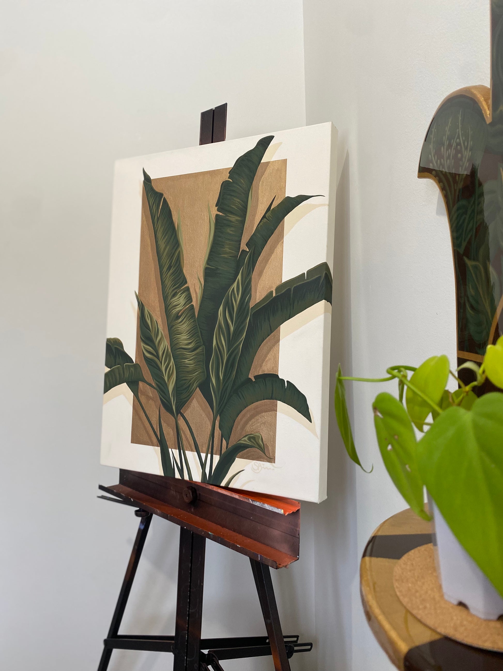 A botanical canvas painting of green tropical leaves on a tan and white background, displayed on a wooden easel in an indoor setting next to a potted plant.